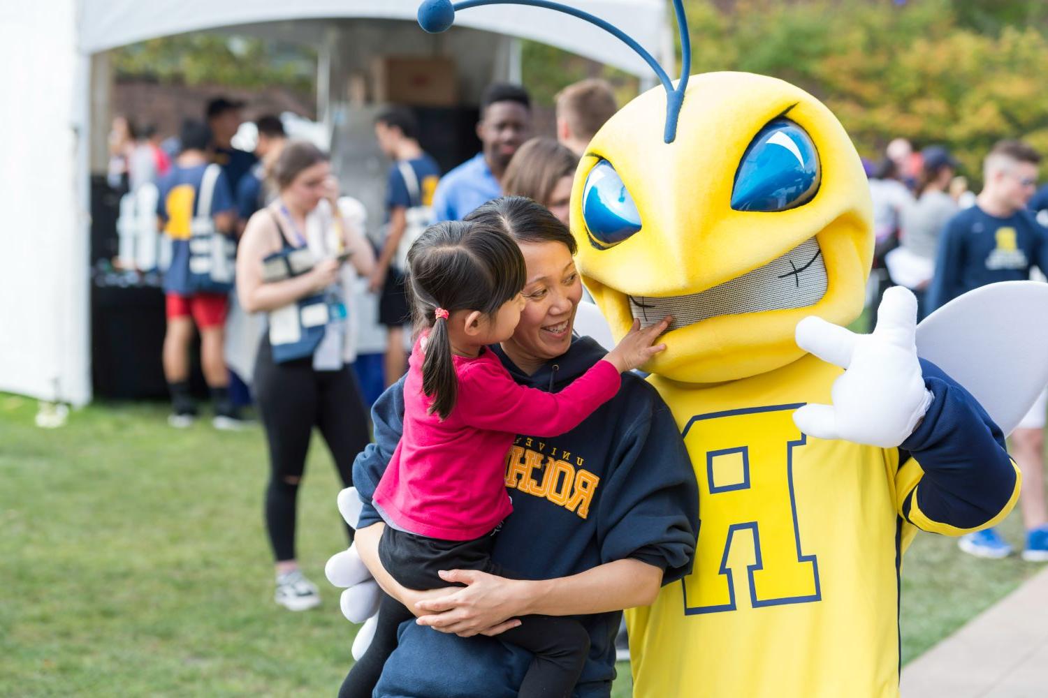 A woman and child embrace a cheerful mascot at a University of Rochester event, showcasing joy and community spirit.