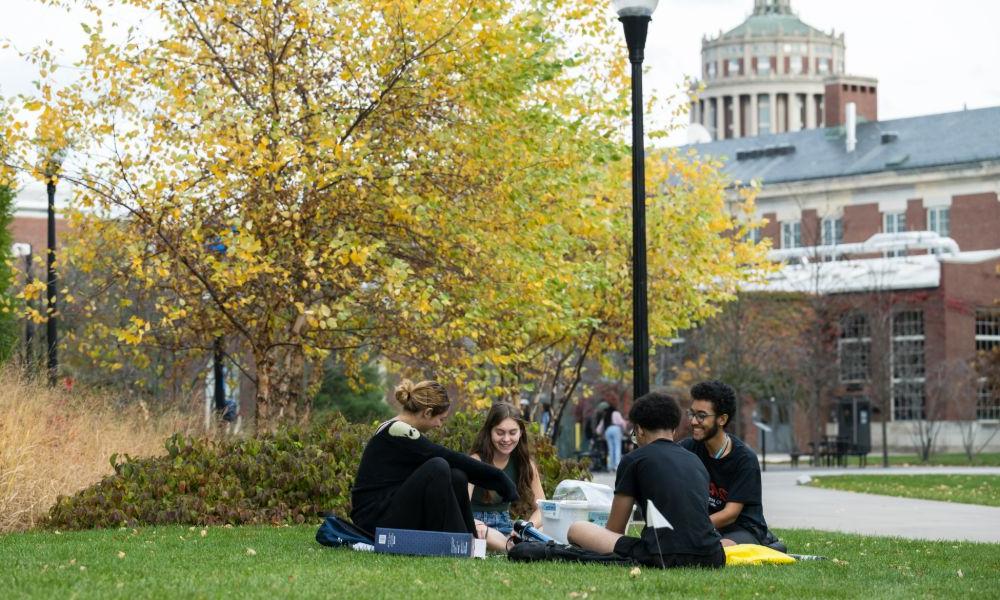 Students relax on the grass in front of the University of Rochester library, enjoying a sunny day outdoors.